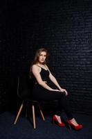 una guapa morena vestida con tacones altos negros y rojos, sentada y posando en una silla en el estudio contra una pared de ladrillo oscuro. retrato de modelo de estudio. foto