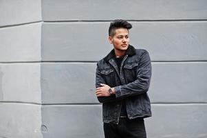 Handsome and fashionable indian man in black jeans jacket posed against gray wall. photo