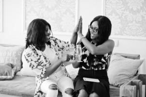 Two african woman friends wear on eyeglasses posed indoor white room and drinking champagne. photo