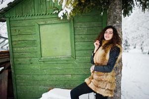 Elegance curly girl in fur coat at snowy forest park agasinst green wooden house at winter. photo