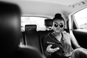 una rica mujer africana de negocios con gafas de sol se sienta en un coche suv con asientos de cuero negro. teléfono móvil a mano. foto