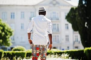 Stylish african american man in white shirt and colored pants with hat and glasses posed outdoor. Black fashionable model boy. photo