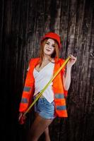 Engineer woman in orange protect helmet and building jacket against wooden background holding tape measure. photo