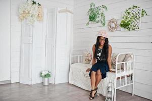 bonita mujer afroamericana con anteojos y sombrero posada en la habitación sentada en un sofá vintage. foto