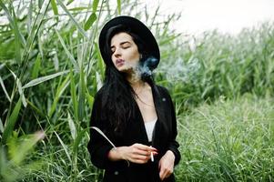 Sensual smoker girl all in black, red lips and hat. Goth dramatic woman smoking on common reed. photo
