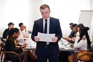 Face of handsome european business man, holding paper with diagram on the background of business peoples multiracial team meeting, sitting in office table. photo