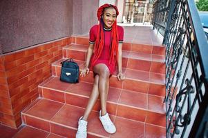 Cute and slim african american girl in red dress with dreadlocks and backpack posed outdoor, sitting on stairs. Stylish black model. photo