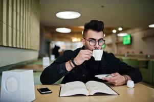 Arab man wear on black jeans jacket and eyeglasses sitting in cafe, read book and drink coffee. Stylish and fashionable arabian model guy. photo