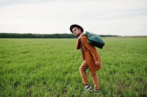 Stylish man in glasses, brown jacket and hat with bag posed on green field. photo