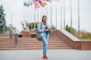 African student female posed with backpack and school items on yard of university, against flags of different countries. photo