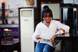 Stylish african american women in white blouse and blue jeans posed at cafe with newspaper. photo