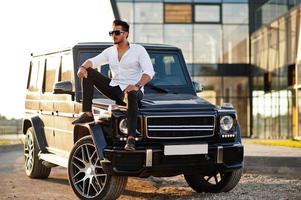 Solid asian man in white shirt and sunglasses posed near black mafia suv car. photo