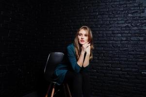 Handsome brunette girl wear on black and green jacket in red high heels, sitting and posing on chair at studio against dark brick wall. Studio model portrait. photo