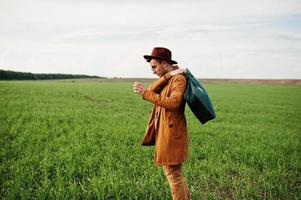 Stylish man in glasses, brown jacket and hat with bag posed on green field. photo