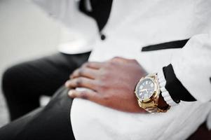 hombre afroamericano elegante y guapo con traje blanco sentado en un banco. foto de primer plano de relojes dorados a mano.