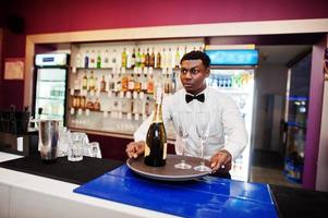 African american bartender at bar holding champagne with glasses on tray. photo