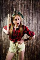 Young funny housewife in checkered shirt and yellow shorts pin up style with kitchen rolling pin on wooden background. photo