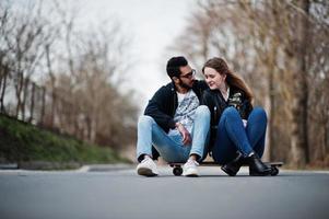 Cool multiracial couple sitting on longboard at road. photo