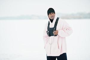 Elegante hombre afroamericano de estilo urbano con capucha rosa posado en un lago congelado en invierno. foto