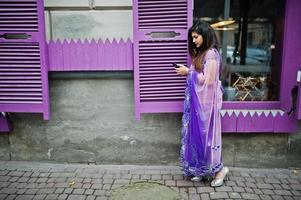 Indian hindu girl at traditional violet saree posed at street against purple windows with mobile phone at hands. photo