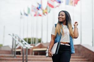 African student female posed with backpack and school items on yard of university, against flags of different countries. photo