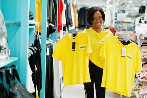 Afican american women in tracksuits shopping at sportswear mall against shelves. She choose yellow t-shirt. Sport store theme. photo