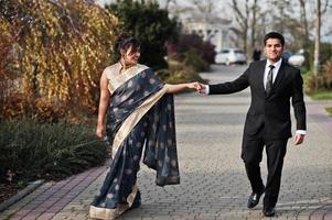 elegante y de moda amigos indios pareja de mujer en sari y hombre en traje caminando al aire libre y tomados de la mano. foto