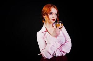 Gorgeous red haired girl in pink blouse and red skirt with glass of wine at hand isolated on black. photo