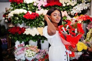 Beautiful african american girl in tender white dress with bouquet flowers in hands standing against floral background in flower shop.Black female florist. photo