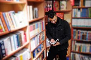 Tall smart arab student man, wear on black jeans jacket and eyeglasses, at library with book at hands. photo