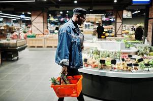 Elegante hombre afroamericano casual con chaqueta de jeans y boina negra sosteniendo una canasta con piñas en la sección orgánica de frutas del supermercado. foto