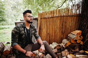 Awesome beautiful tall ararbian beard macho man in glasses and military jacket posed outdoor, sitting on stumps. photo