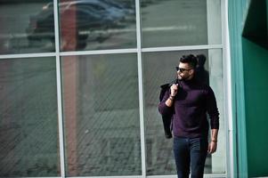 Fashion rich beard Arab man wear on turtle neck and sunglasses posed against large windows of modern building with jacket on hand. Stylish, succesful and fashionable arabian model guy. photo