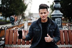 Handsome and fashionable indian man in black jeans jacket posed outdoor, sitting on bench with mobile phone at hand, showing thumb up. photo