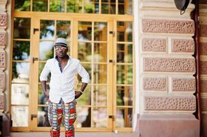 elegante hombre afroamericano con camisa blanca y pantalones de colores con sombrero y gafas al aire libre. chico modelo de moda negro. foto