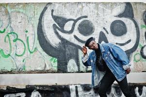 hombre afroamericano con chaqueta de jeans, boina y anteojos contra la pared de graffiti con calavera. foto