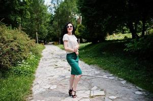 Brunette girl in green skirt and white blouse with sunglasses posed at park. photo