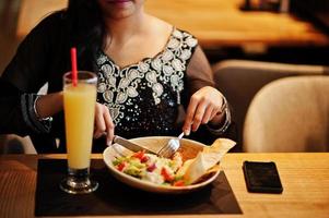 linda chica india vestida con sari negro posó en el restaurante, sentada a la mesa con jugo y ensalada. foto