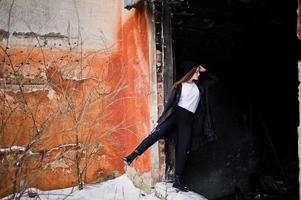 Fashionable long legs brunette model in long black cloak posed outdoor at winter day against old grunge wall. photo