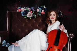 Pretty young gilrl musician in white dress with double bass sitting on brown vintage sofa. photo
