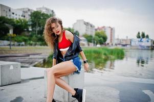 Sexy curly model girl in red top, jeans denim shorts, leather jacket and sneakers posed at stone cubes. photo