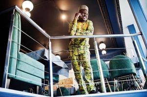 Friendly afro man in traditional yellow clothes with cap at restaurant near the handrail speaking at mobile phone. photo