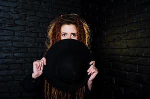 Studio shoot of girl in black with dreads and hat on brick background. photo