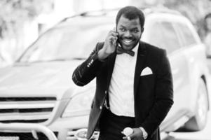 Handsome fashionable african american man in formal wear and bow tie with walking stick and mobile phone stay against business suv car. photo