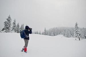 Man tourist photographer with backpack, at mountain with pine trees covered by snow. Beautiful winter landscapes. Frost nature. photo