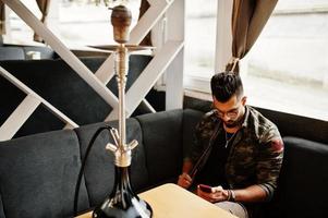 Stylish beard arabian man in glasses and military jacket smoking hookah at street bar. Arab model having rest and looking on phone. photo