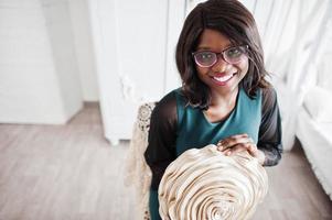 Pretty african american woman in eyeglasses posed in room, sitting on chair with decorative flower in hands. photo