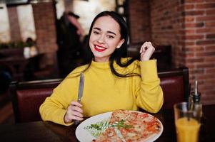 chica morena divertida en suéter amarillo comiendo pizza en el restaurante. foto