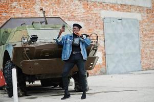 African american man in jeans jacket, beret and eyeglasses, with cigar posed against btr military armored vehicle, and making selfie on mobile phone. photo