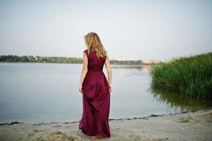 Back of blonde sensual barefoot woman in red marsala dress posing against lake with reeds. photo